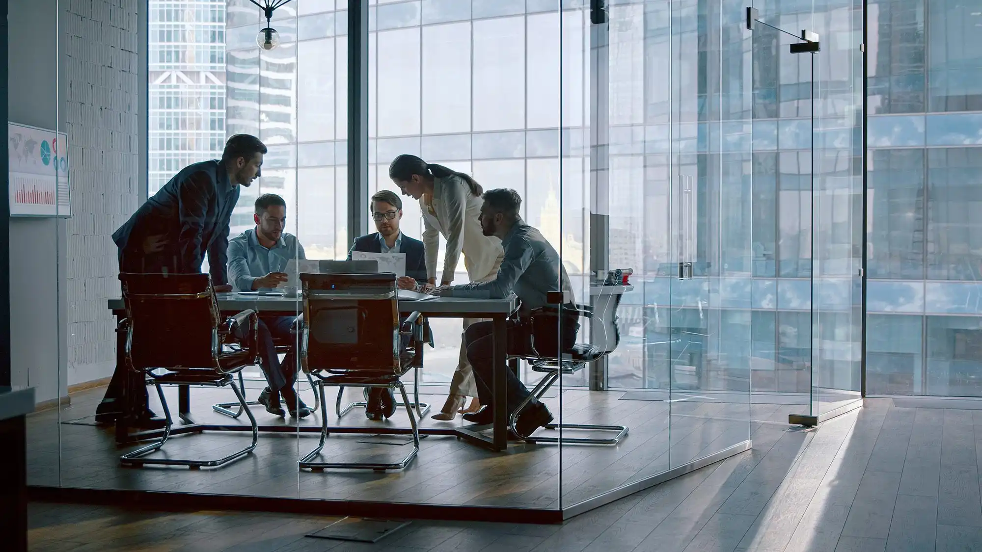 Five office workers huddle around a conference table, examining a laptop and papers while one leans in to point