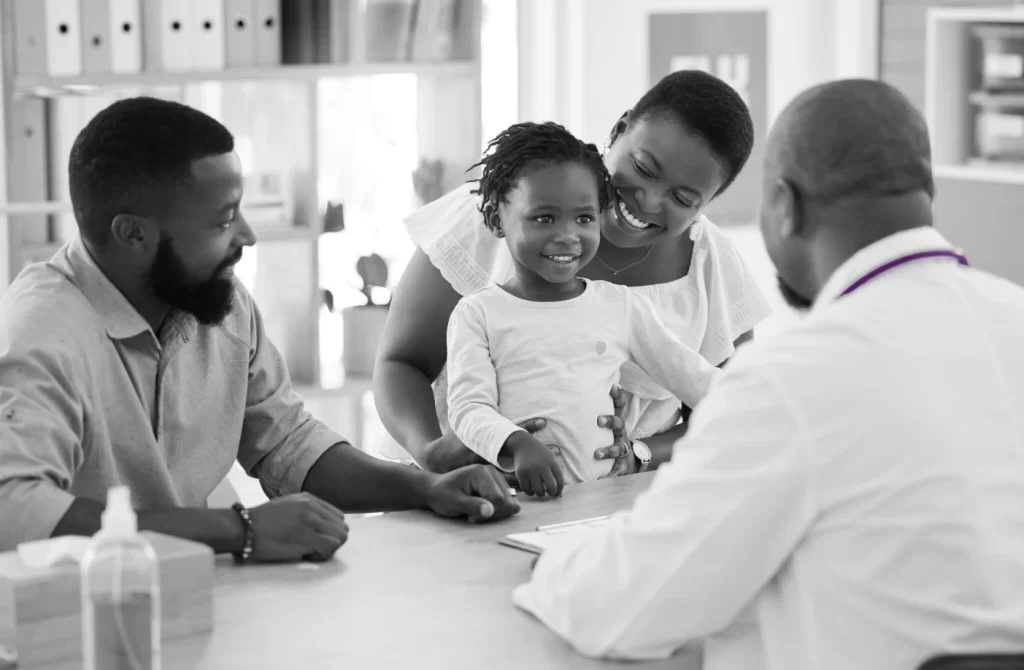 Young child smiles, sitting on a woman's lap while a man and a doctor converse across a desk in a bright medical office.