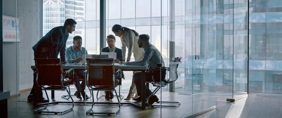 Five office workers huddle around a conference table, examining a laptop and papers while one leans in to point